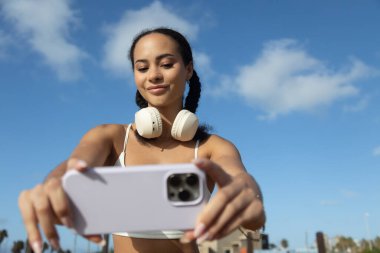 Mavi gökyüzü arka planlı cep telefonuyla selfie çeken genç Latin kadının portresi.