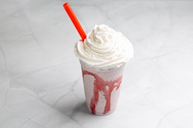 Kremalı çilekli milkshake kremayla dolup taşıyor, parlak mermer yüzeyde dinleniyor, kırmızı pipet ortaya çıkıyor.