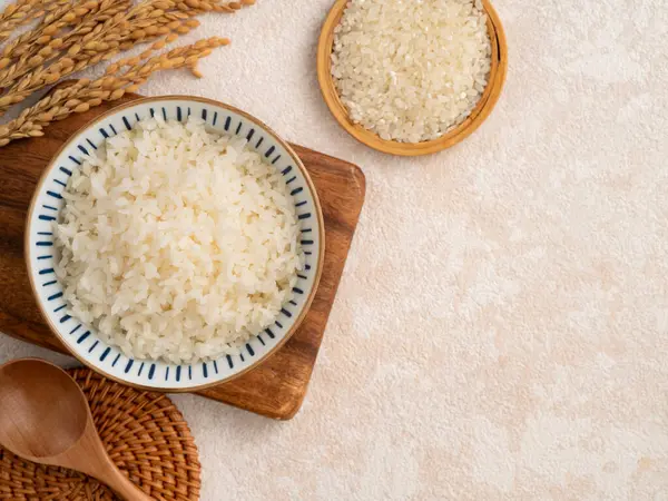 Bir kase taze pişmiş beyaz pirinç, yumuşak, kabarık ve beyaz masa arkasında hafifçe parıltılı..