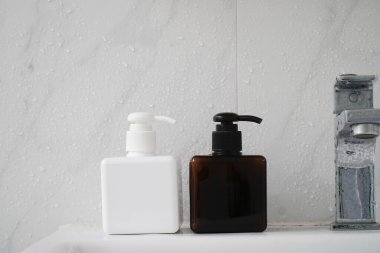Two square-shaped bottles, one white and one brown, with black pumps, sit on a wet sink in front of a white-tiled wall. clipart