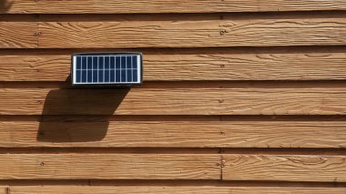 Küçük güneş paneli ahşap duvara monte edilir, gölge bırakır. Tahta doğal, dokulu bir görünüme sahiptir ve panel güneş ışığını etkili bir şekilde yakalamak için konumlandırılmıştır.