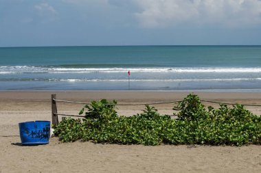 Huzurlu Bali sahili, yumuşak dalgalar, kırmızı bir güvenlik bayrağı ve yemyeşil deniz kenarındaki huzurlu anları davet ediyor.