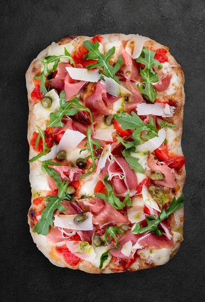 Sorrentina pizza with prosciutto, arugula, capers, pelati sauce, pesto. Roman pizza rectangular on dark background