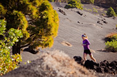 Trail running athlete training on a sunny day in a volcanic landscape of black lava sand with green trees and a path marked with stones. The woman is dressed in sneakers, leggings and long tight t-shirt and a headband.