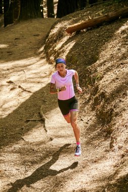 Ormanın içindeki toprak yolda koşan sporcu pembe tişört, mavi kurdele, siyah tayt ve mor spor ayakkabı giyiyor..