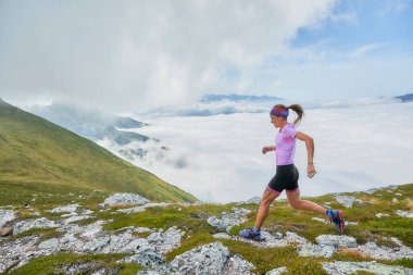 Dağa tırmanan bir kadın. Kayalar ve derin yeşil çimlerle. Arkasında beyaz bulutlardan oluşan bir deniz var..