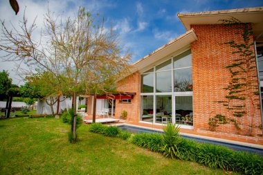 Çimenlerle, ağaçlarla ve bitkilerle çevrili yüzme havuzlu kır evi, güneşli bir yaz gününde ön planda bir ağaç, mavi gökyüzü ve beyaz bulutlar..