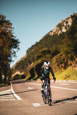 Güneşli bir günde, yalnız bir yolda siyah bir forma ve önlük şort ve beyaz bir kaskla bisiklet eğitimi alan bir kadın.. 