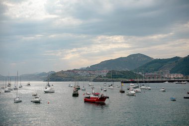 Hareketli bir kıyı kasabasında, canlı balıkçı tekneleri şehrin renkli binaları ve arka planda yeşil tepelerle birlikte limanda dinleniyor. Sakin su tekneleri yansıtıyor ve bulutlu bir gökyüzünün altında sakin bir atmosfer yaratıyor. Sahne yakalar