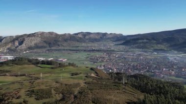 Bir insansız hava aracı, Los Corrales de Buelna kasabasını bir dağın tepesinden ortaya çıkararak, açık bir gökyüzünün altındaki yeşil tepelerin ve dağların arasına yerleşmiş kentsel ve kırsal manzaraların karışımını sergiliyor..