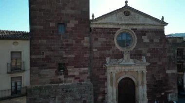 Santa Maria de Prades 'in çan kulesini ortaya çıkarmak için bir dron yükselir, sabahın erken saatlerinde güneş tarafından çerçevelenir, köyün çatıları ve uzak tepeleri arka planda ortaya çıkar, sakin ve zamansız bir sahne yaratır..