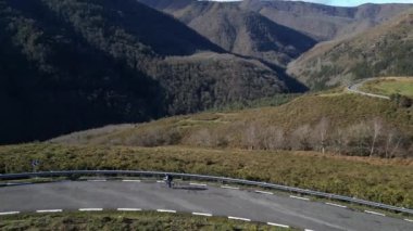 Bir bisikletçi, açık mavi gökyüzü altında yemyeşil ve dramatik yamaçlarla çevrili, dolambaçlı dağ vadilerinde dolambaçlı bir yolda gezinerek açık hava macerasının özgürlüğünü yakalıyor..