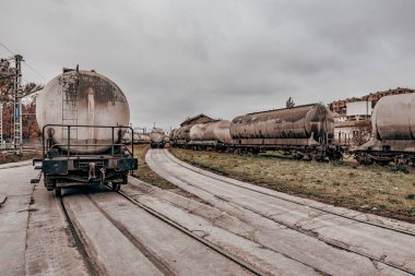 Endüstriyel Demiryolu Tersanesinde Yük ve Yolcu Trenleri. Bulutlu bir günde yük vagonları ve yolcu trenleriyle sanayi bölgesi. Paslı tank vagonları, elektrik hatları ve raylar cesur ve faydacı bir atmosferi çağrıştırıyor..