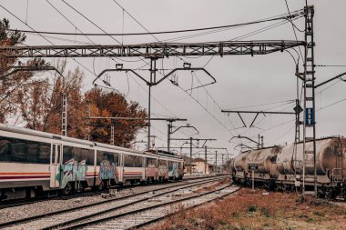 Endüstriyel Demiryolu Tersanesinde Yük ve Yolcu Trenleri. Bulutlu bir günde yük vagonları ve yolcu trenleriyle sanayi bölgesi. Paslı tank vagonları, elektrik hatları ve raylar cesur ve faydacı bir atmosferi çağrıştırıyor..