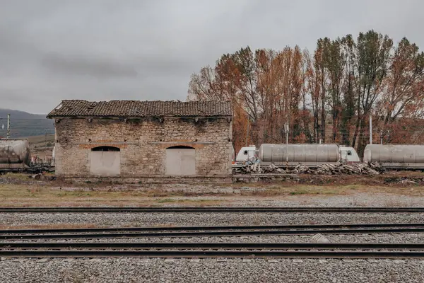 Demiryolu raylarının oradaki terk edilmiş taş bina. Pencereleri tahta kaplı eski bir taş bina demiryolu raylarının yanında duruyor. Sonbahar ağaçları ve yük trenleriyle çevrili, nostaljik ve terk edilmiş bir atmosfer yayar..