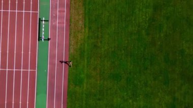 İHA, yüksek bir hava perspektifinden yakalanmış bir sporcuyu pistin kenarında ve çimenli alanda takip ediyor..