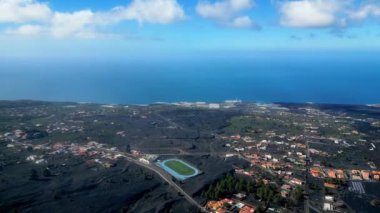 La Palma adasının havadan görünüşü, kararmış manzaralar, dağınık yerleşim alanları ve açık mavi gökyüzünün altındaki Atlantik Okyanusu ile birlikte volkanik bir patlamanın sonrasını gösteriyor..