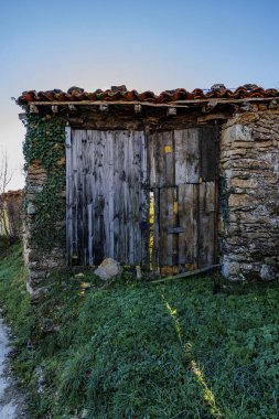 Yıpranmış ahşap bir kapı. Etrafı sarmaşık kaplı taş duvarlarla çevrili. Sessiz kırsal bir ortamda.