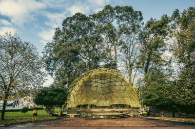 Parlak mavi gökyüzünün altında ağaçlarla çevrili canlı sarı kubbeleri ve kırmızı bankları olan bir açık hava amfi tiyatrosu. Kültürel ve doğal uyumu yakalar.