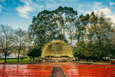 An outdoor amphitheater featuring a vibrant yellow dome and red benches, surrounded by trees under a bright blue sky. Captures cultural and natural harmony. clipart