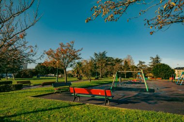 A vibrant playground surrounded by benches and trees in a sunny park. Captures a serene outdoor setting for relaxation and recreation. clipart