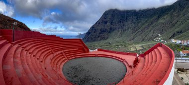 Traditional Canarian wrestling arena in El Hierro, Spain, with red stands and a stunning mountain and ocean backdrop. A unique cultural landmark blending sports and breathtaking scenery. clipart