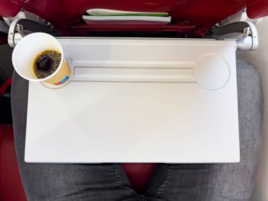 Passenger in economy class with a coffee cup on an airplane tray table. The compact space, red seat, and travel essentials capture the inflight experience. clipart