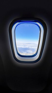 Parlak mavi gökyüzünü ve kabarık bulutları gösteren bir uçak penceresinden hava manzarası. Karanlık kabin ile gökyüzü arasındaki zıtlık seyahat deneyimini güçlendirir..