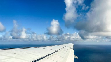 Aerial view of an airplane wing flying over the ocean with scattered clouds. The blue sky and horizon create a stunning travel perspective from above. clipart