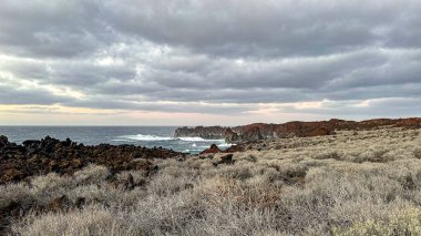 Güçlü okyanus dalgalarının kayalıklara çarptığı engebeli volkanik kıyı şeridi. Dramatik manzara, karanlık kayalar ve bulutlu gökyüzü vahşi ve uzak bir atmosfer yaratır..