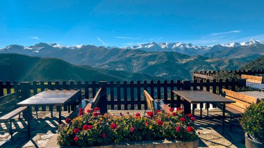Ahşap masaları ve çiçek saksıları olan rahat bir dağ terası. Açık mavi gökyüzünün altında, karla kaplı tepelerin büyüleyici panoramik manzarasına bakıyor. Rahatlamak ve manzaralı yemek için mükemmel..