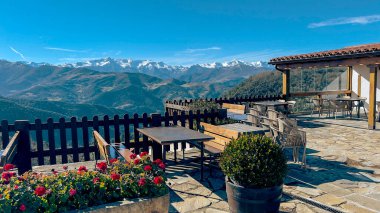 A cozy mountain terrace with wooden tables and flowerpots overlooking a stunning panoramic view of snow-covered peaks under a clear blue sky. Perfect for relaxation and scenic dining. clipart