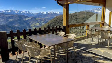 Ahşap masaları ve çiçek saksıları olan rahat bir dağ terası. Açık mavi gökyüzünün altında, karla kaplı tepelerin büyüleyici panoramik manzarasına bakıyor. Rahatlamak ve manzaralı yemek için mükemmel..