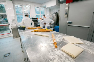 Paslanmaz çelik bir masa üzerinde merdane, un ve hamurlu bir fırın mutfağı. Pasta hazırlamada birlikte çalışan beyaz üniformalı profesyonel fırıncılar..