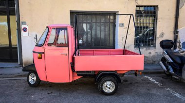 Üç tekerlekli pembe bir araç, arka planda motosiklet olan bir caddeye park edilmiş..