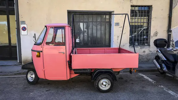 Üç tekerlekli pembe bir araç, arka planda motosiklet olan bir caddeye park edilmiş..