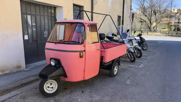 Üç tekerlekli pembe bir araç, arka planda motosiklet olan bir caddeye park edilmiş..