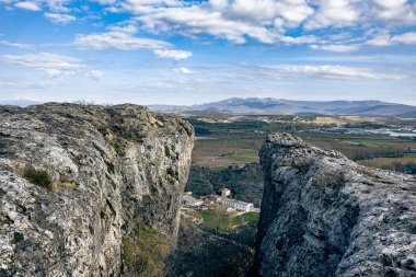 Yüksek kayalık kayalıklardan, etrafı tarlalarla çevrili küçük bir köye ve dağınık bulutlu mavi gökyüzünün altındaki uzak dağlara bakıyor..