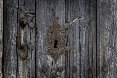 Eski yıpranmış ahşap bir kapıya yakın çekim. Süslü paslı metal kilidi ve desenli tahılları var. Kaliteli, kırsal ayrıntı güçlü karakter ve patinaj ile.