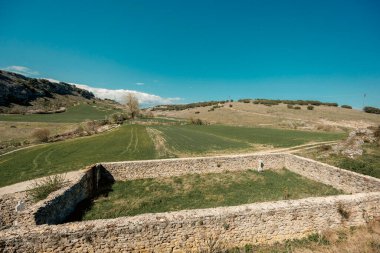 Parlak mavi bir gökyüzünün altında yeşil tarlaları, tepeleri ve toprak yolları olan kırsal bir vadide duvarlı bir taşın manzarası. Huzurlu ve zamansız kırsal alan.