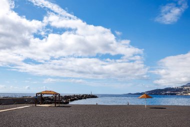 Turuncu şemsiyeli geniş kumlu sahil ve açık mavi gökyüzünün altında boş kıyı şeridi. Yazın başlarındaki sessiz beklentileri simgeliyor..