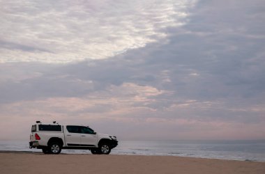 Beyaz off-road arabası Atlantik okyanusunun kıyısında duruyor. Parlak dramatik bir günbatımında.