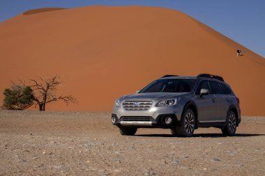 Sıcak ve güneşli bir günde, kızıl bir kum tepeciğinin yanında gri bir Subaru. 