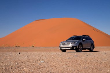 Sıcak ve güneşli bir günde, kızıl bir kum tepeciğinin yanında gri bir Subaru. 