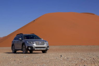 Sıcak ve güneşli bir günde, kızıl bir kum tepeciğinin yanında gri bir Subaru. 