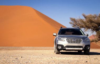 Sıcak ve güneşli bir günde, kızıl bir kum tepeciğinin yanında gri bir Subaru. 