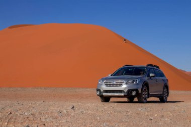 Sıcak ve güneşli bir günde, kızıl bir kum tepeciğinin yanında gri bir Subaru. 