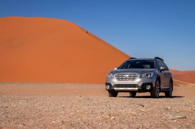 Sıcak ve güneşli bir günde, kızıl bir kum tepeciğinin yanında gri bir Subaru. 