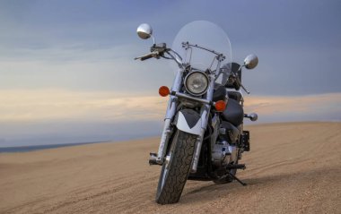 Namib Çölü 'nün kumlu yüzeyinde dramatik bulutlu bir gökyüzüne karşı tek başına duran bir motosiklet. Günbatımı ışığı, doğayla makine arasındaki zıtlığı vurgular..