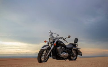 Namib Çölü 'nün kumlu yüzeyinde dramatik bulutlu bir gökyüzüne karşı tek başına duran bir motosiklet. Günbatımı ışığı, doğayla makine arasındaki zıtlığı vurgular..